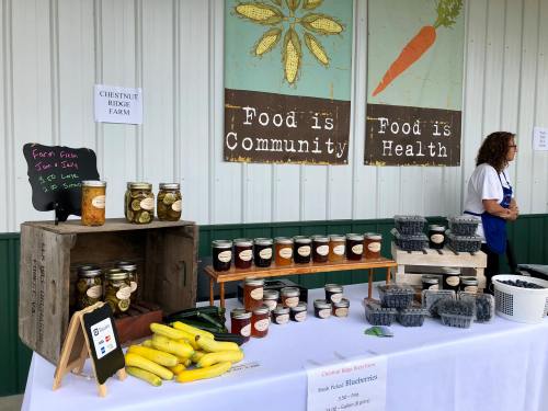 A farm stand displays jars of preserves, fresh produce, and a vendor in front of colorful signs about food and community.