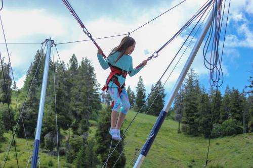 A young girl in a harness jumps on a bungee trampoline, surrounded by trees and a blue sky.