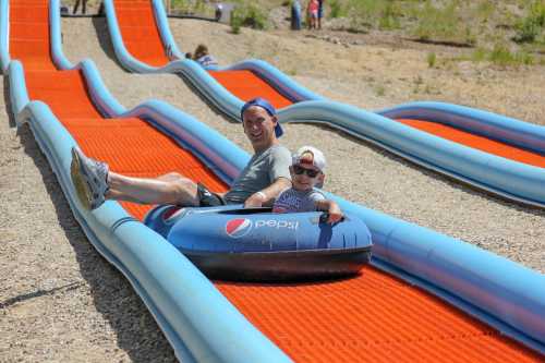 A man and a child sit in a blue inflatable tube on a colorful water slide, smiling as they prepare to slide down.