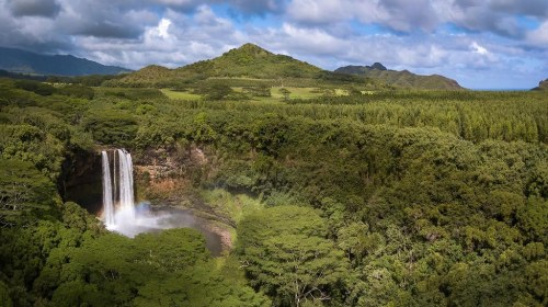 A stunning waterfall cascades into a lush green valley, surrounded by mountains and a blue sky with clouds.