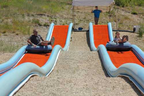 Two people on inflatable tubes slide down orange and blue water slides, with a tent and a person in the background.