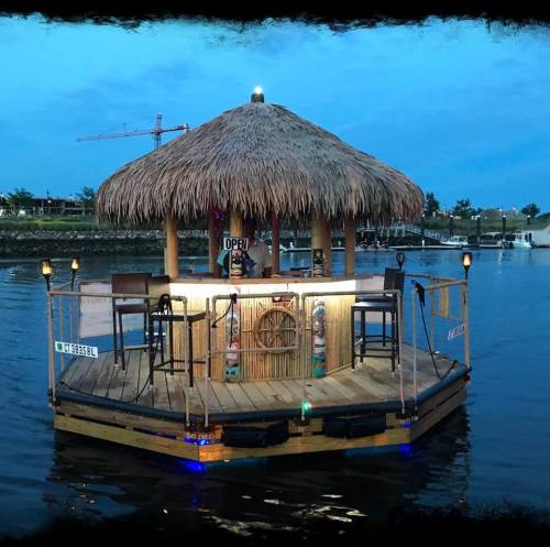 A floating tiki bar with a thatched roof, surrounded by water, featuring stools and lights at dusk.