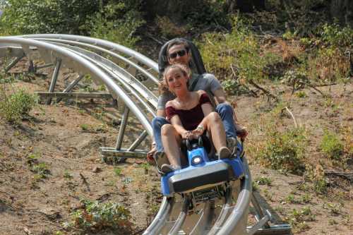 A man and woman enjoy a ride on a roller coaster, smiling as they descend a track through a wooded area.