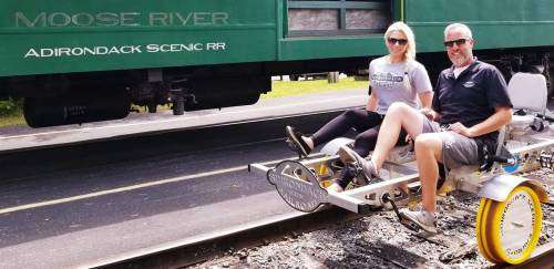 A man and woman sit on a rail bike beside a green train, enjoying a scenic ride along the Moose River.