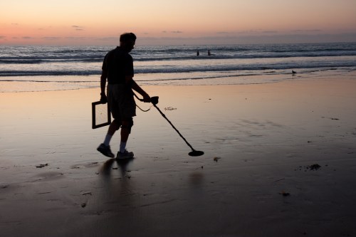 A person using a metal detector walks along the beach at sunset, with gentle waves in the background.