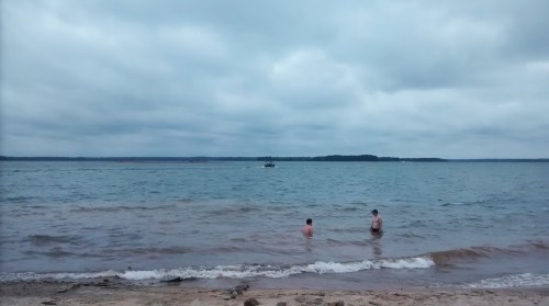 Two people wade in a calm lake under a cloudy sky, with a boat visible in the distance.