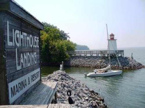 A sailboat approaches a lighthouse near a marina, with rocky shores and greenery in the background.