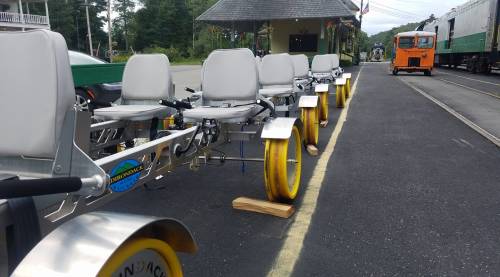 Row of pedal-powered rail bikes lined up on a track, with a vintage train in the background.