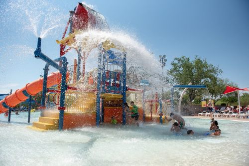 A colorful water play structure with slides and water sprays, surrounded by children playing in a sunny pool area.