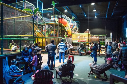 A busy indoor play area with children playing on equipment and families sitting on benches, enjoying the space.
