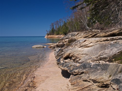 Rocky shoreline with clear water and trees under a blue sky. Calm lake reflects the serene landscape.