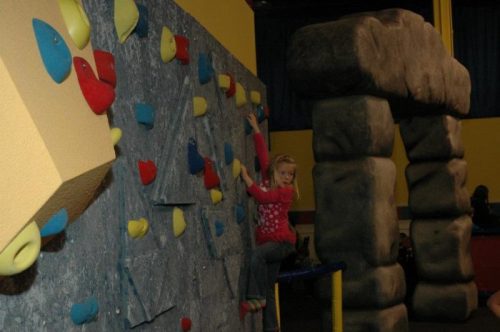 A child climbs a colorful indoor rock wall with a stone structure nearby.
