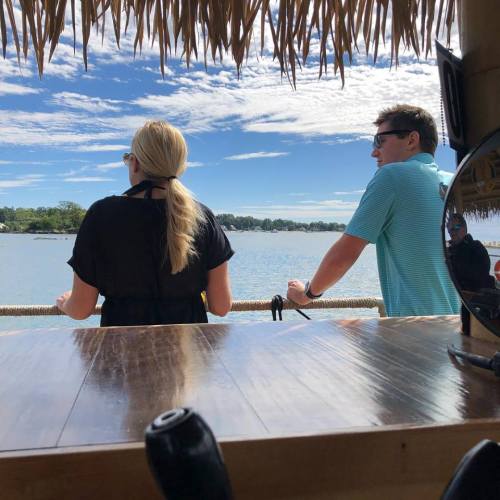 Two people stand by a waterfront, looking out at the water under a thatched roof, with a clear blue sky above.
