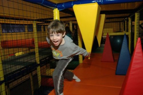 A smiling boy runs through a colorful indoor play structure with soft shapes and climbing areas.