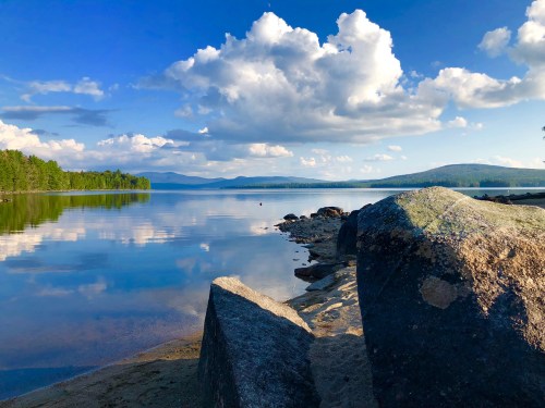 A serene lake scene with calm water reflecting clouds and trees, framed by large rocks on the shore.