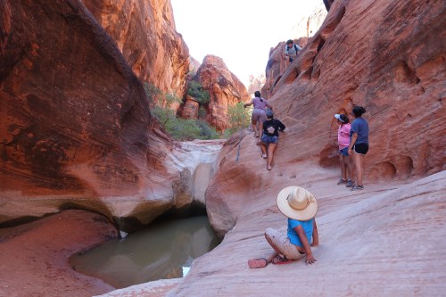 A group of people exploring a narrow canyon with red rock formations and a small pool of water.