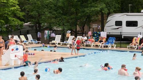 A busy pool scene with children swimming, adults lounging on chairs, and towels hanging nearby under trees.