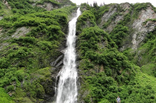 A tall waterfall cascades down a lush, green mountainside, with a person standing at the base for scale.