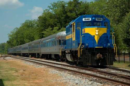 A blue and yellow locomotive pulls a silver passenger train along a tree-lined railway track on a sunny day.