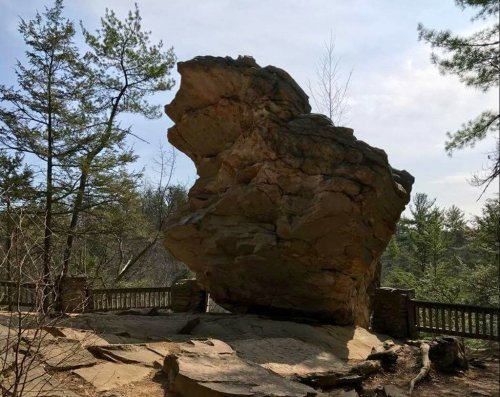 A large, tilted rock formation surrounded by trees and a wooden railing under a clear sky.