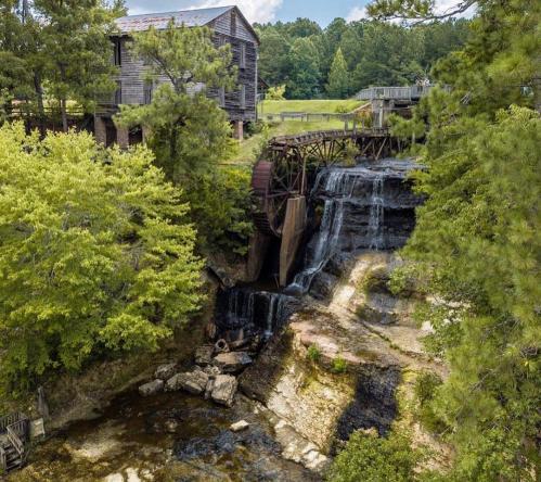 A scenic view of a waterfall cascading over rocks, with a historic wooden mill and lush greenery in the background.