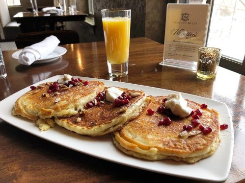 Three fluffy pancakes topped with pomegranate seeds and dollops of cream, served with a glass of orange juice.