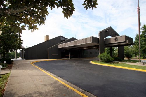 Modern building with a dark exterior, featuring a covered walkway and surrounded by greenery and a clear sky.