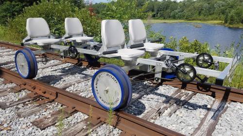 A four-seat rail cart on a gravel track near a lake, surrounded by greenery.