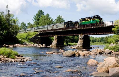 A train crosses a bridge over a river, surrounded by lush greenery and rocky banks under a blue sky.