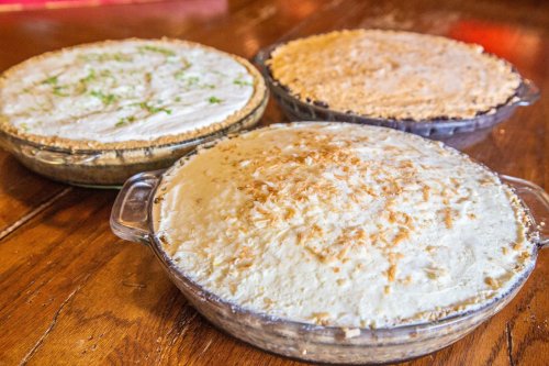 Three delicious pies on a wooden table: a coconut cream pie, a chocolate pie, and a key lime pie.
