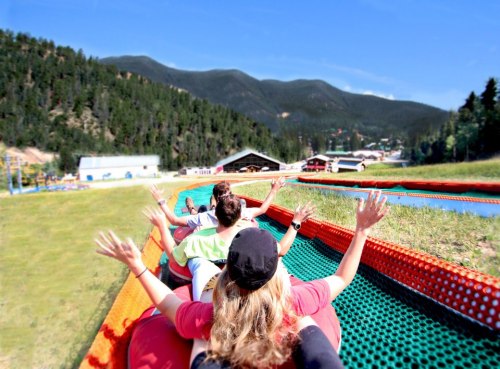 A group of people enjoying a summer slide ride down a grassy hill, surrounded by mountains and trees.