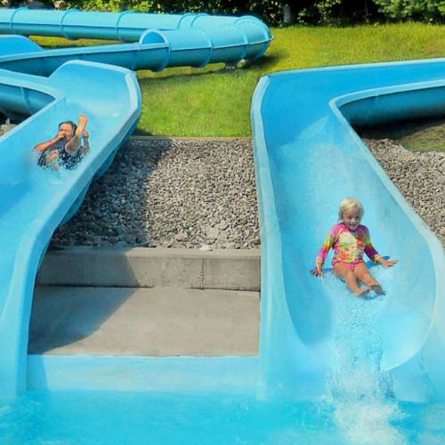 Two children slide down bright blue water slides into a pool, surrounded by green grass and gravel.