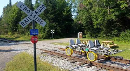 A railroad crossing sign next to a small rail cart on tracks, surrounded by trees and a picnic area in the background.