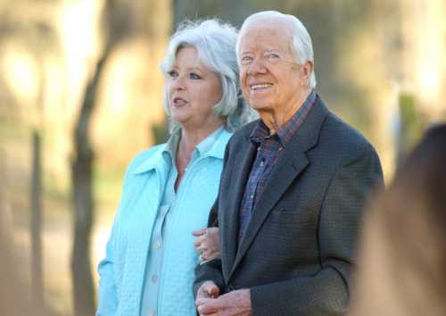 A smiling elderly couple stands together outdoors, with trees in the background. The woman wears a light blue jacket.