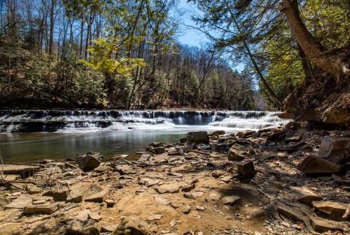 A serene river scene with a gentle waterfall, surrounded by lush trees and rocky terrain under a clear blue sky.
