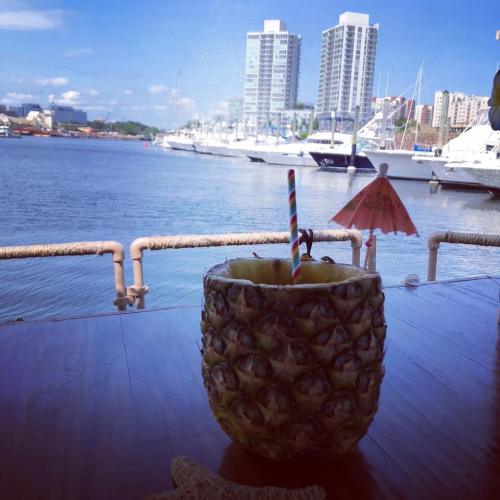 A pineapple drink with a straw sits on a table by the water, with boats and tall buildings in the background.