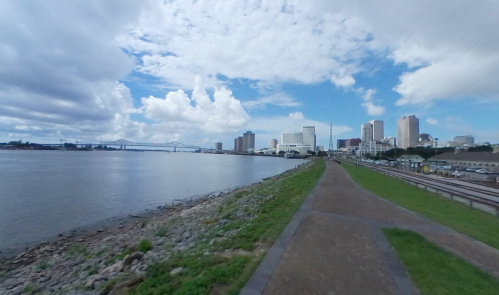 A scenic view of a riverbank with a pathway, city skyline, and cloudy blue sky in the background.