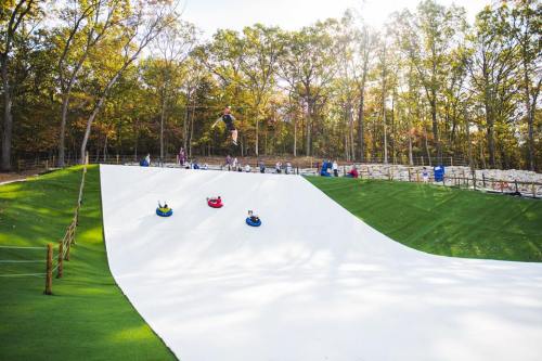 A snowy hill with people sledding down on colorful tubes, surrounded by trees and a sunny sky.