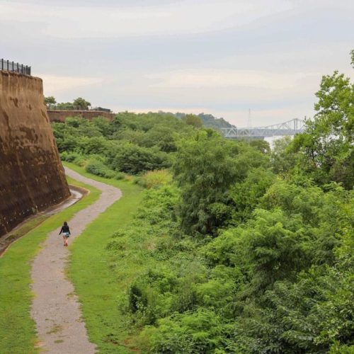 A person walks along a path beside a large, weathered wall, surrounded by lush greenery and a river in the distance.