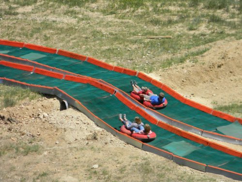 Two people on inner tubes slide down a grassy, winding slope with orange safety barriers.