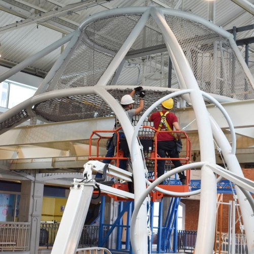 Two workers on a lift inspect a large metal structure indoors, wearing safety helmets and harnesses.