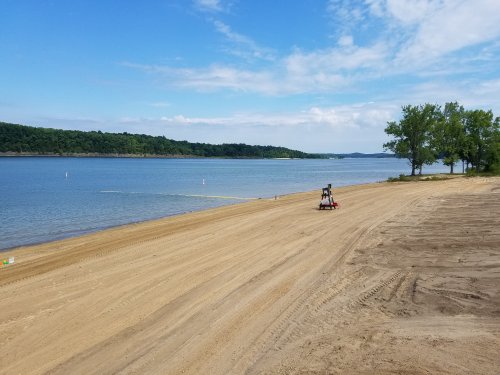 A sandy beach by a calm lake, with a lone beach chair and trees in the background under a partly cloudy sky.