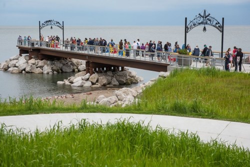 A crowded pier extends over water, surrounded by rocks and green grass, under a cloudy sky.