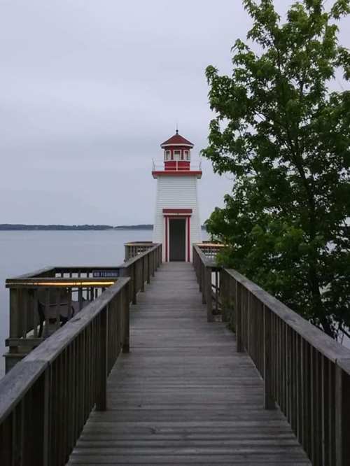 A wooden walkway leads to a white lighthouse with red accents, surrounded by calm water and greenery.