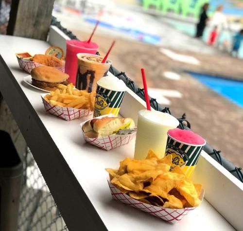 A variety of colorful fast food items on a table, including burgers, fries, drinks, and desserts, near a pool.