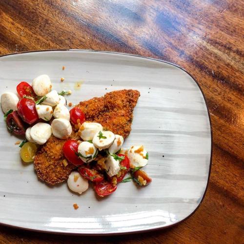 A plate with crispy fried fish topped with cherry tomatoes, mozzarella balls, and herbs on a wooden table.