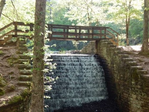 A serene waterfall flows beneath a wooden bridge, surrounded by lush green trees and natural stone steps.