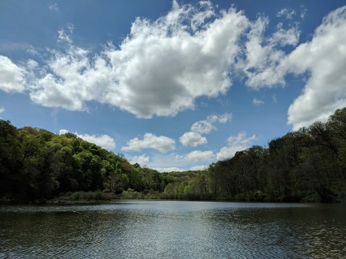 A tranquil lake is framed by green trees under a blue sky with white clouds.