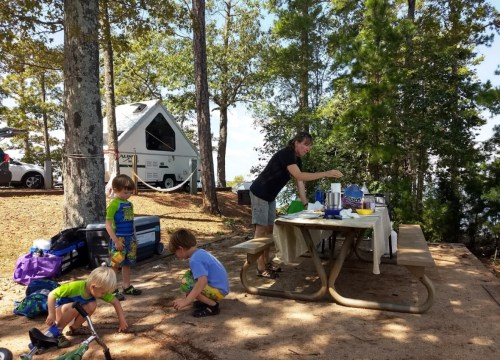 A family camping scene with children playing on the ground while an adult prepares food at a picnic table.