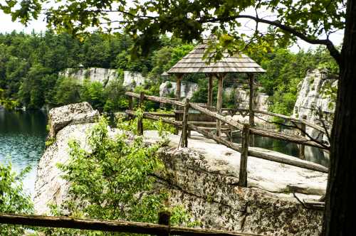 A wooden gazebo on a rocky ledge overlooking a serene lake surrounded by lush greenery and cliffs.